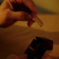 A hand holds a small glass perfume vial labeled 'Rosemary Odor' above an open black spray bottle on a wooden surface, lit by warm yellow light.