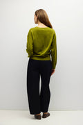 A woman is shown from the back wearing a textured olive green sweater and dark wide-leg trousers, standing against a plain light gray studio background.