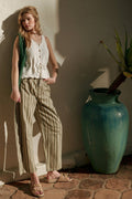 Woman in a white sleeveless top and striped pants standing next to a large green vase on a stone floor.
