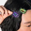 A close-up lifestyle shot of a person with dark hair wearing two metallic beetle-shaped hair clips, one purple and one green with red accents, held by a hand.