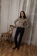 A woman wearing a beige and black vertically striped long-sleeve top, dark navy pants, and black boots stands in a room with sheer white curtains and a wooden floor, posing next to a wooden chair.