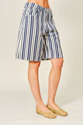 A person wearing knee-length vertical striped shorts in navy blue and off-white, paired with woven tan flat shoes, photographed against a plain light beige studio background.