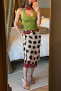 A woman takes a mirror selfie in a bedroom setting, wearing a green tank top, a beige wrap-style midi skirt with large black polka dots and red trim, and red strappy heels.