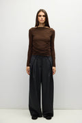 A model stands against a light gray studio background wearing a brown off-the-shoulder long-sleeve top and black wide-leg trousers.