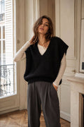 A woman stands indoors wearing a black sleeveless knit V-neck sweater vest over a cream long-sleeve top, paired with dark gray trousers. The setting features a warm beige wall, a window with shutters, and a stone fireplace.