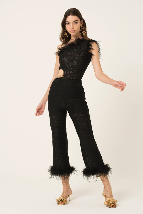 A model wearing a black lace and feather one-shoulder jumpsuit with a side cutout and matching feather-trimmed ankle cuffs, paired with gold platform sandals, against a plain off-white studio background.