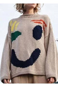 A person wearing a chunky knit sweater with a large abstract smiley face design in beige, navy, green, yellow, and orange, photographed outdoors against a pale sky with birds in the background.