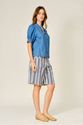 A woman modeling a blue denim short-sleeve button-up shirt and blue-and-white vertically striped shorts, paired with tan woven flat shoes, against a plain light beige studio background.