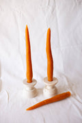 Two tall orange candles in white holders with a carrot on a white background