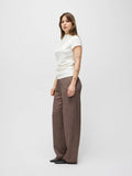 A woman stands in profile wearing a white short-sleeve knit top and brown wide-leg trousers against a plain light gray studio background.