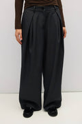A person wearing black wide-leg pleated trousers with a matching black belt, paired with a brown long-sleeve top and black loafers, shown from the waist down against a plain light gray studio background.