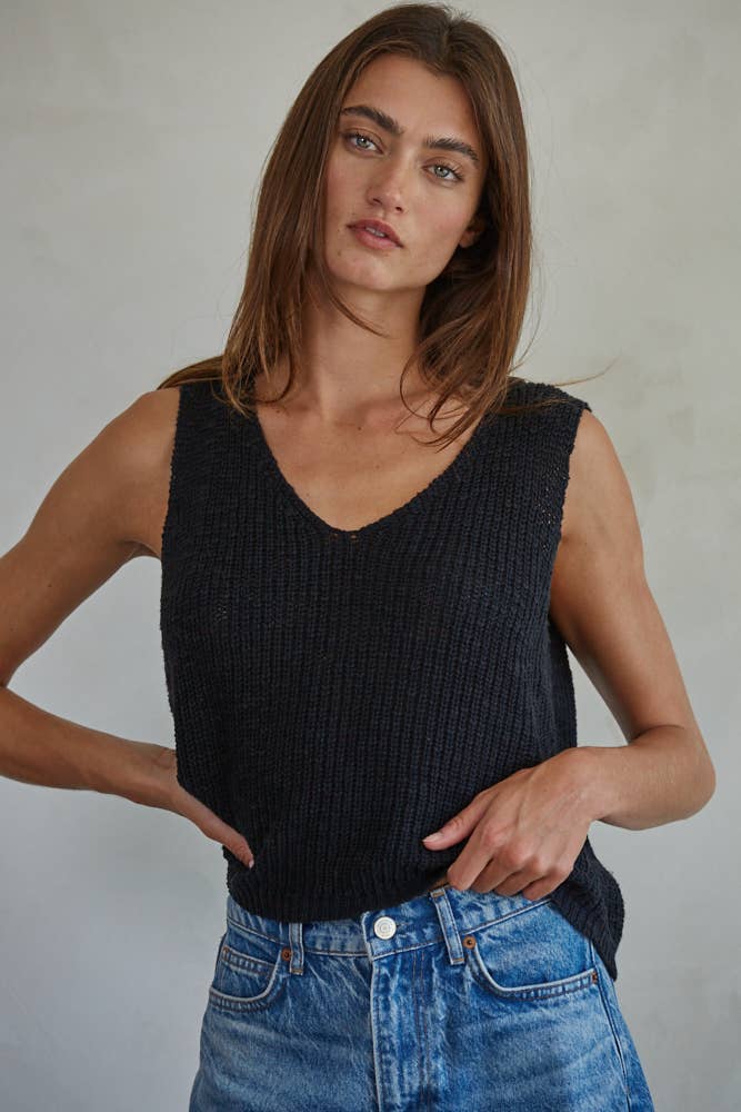 Woman wearing a black sleeveless knitted top and blue jeans against a plain background