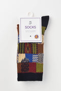 A pair of multicolor patchwork knit socks hanging from a white paper hanger with a product label that reads 'SOCKS'. The socks feature a patchwork design with various geometric and textured patterns in green, blue, red, brown, and beige, finished with solid black ribbed cuffs.
