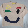 A beige knit sweater laid flat on a light blue-gray surface, featuring a stylized face design in navy, green, yellow, black, and red.