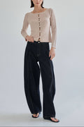 A woman stands against a light gray studio background wearing a beige ribbed long-sleeve top with front buttons and dark wide-leg jeans.