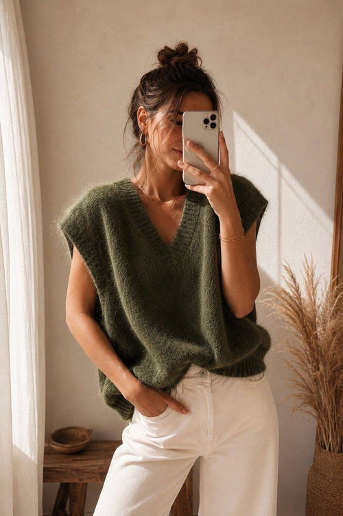A woman takes a mirror selfie wearing a fuzzy olive green sleeveless V-neck sweater and white pants in a warm, sunlit room.