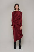 A woman modeling a long-sleeved, gathered midi dress in deep burgundy with black boots, standing against a neutral light gray studio background.