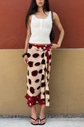 A woman models a wrap skirt featuring a beige base with large dark brown spots and red side panels with white polka dots, styled with a white tank top and black sandals against a two-toned wall.
