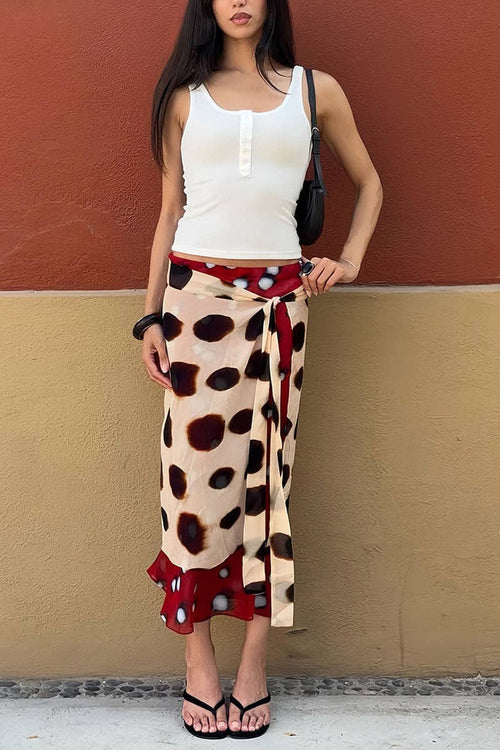 A woman models a wrap skirt featuring a beige base with large dark brown spots and red side panels with white polka dots, styled with a white tank top and black sandals against a two-toned wall.
