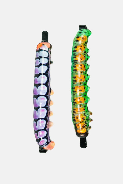 Two decorative hair claw clips shaped like caterpillars, shown against a light gray background. The top clip is purple and white with peach accents, and the bottom clip is green and orange with black spots.
