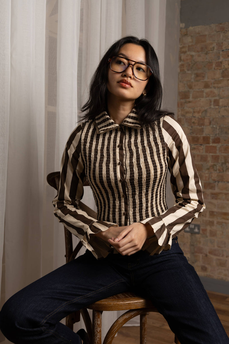 A woman wearing a brown and beige vertically striped long-sleeve button-up shirt and dark blue jeans, sitting on a wooden stool in an indoor setting with a brick wall and white curtains.