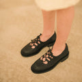Black flat shoes with cut-out design worn by a person on a beige carpet.