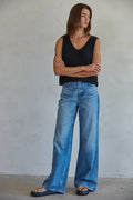 Person wearing a black sleeveless top and blue jeans standing against a plain wall.
