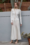 Woman wearing a white outfit with a patterned skirt standing outdoors against a wooden fence.