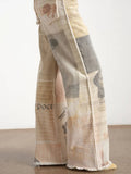 A close-up of a person's legs wearing wide-leg trousers made from a distressed, patchwork-style newspaper print in beige, cream, gray, and brown tones.