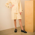 Person wearing a light yellow outfit with black shoes standing against a beige wall.