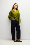 A woman stands against a plain light gray studio wall wearing a long-sleeve olive green V-neck knit sweater, dark navy wide-leg trousers, and brown loafers.