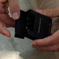 A person's hands holding an open black box of elemeter discovery perfume oil, showing the small round bottles inside, against a textured gray background.