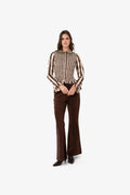 A woman modeling a brown and beige vertically striped long-sleeve blouse with ruffled cuffs, paired with dark brown flared trousers and black shoes, against a plain white studio background.