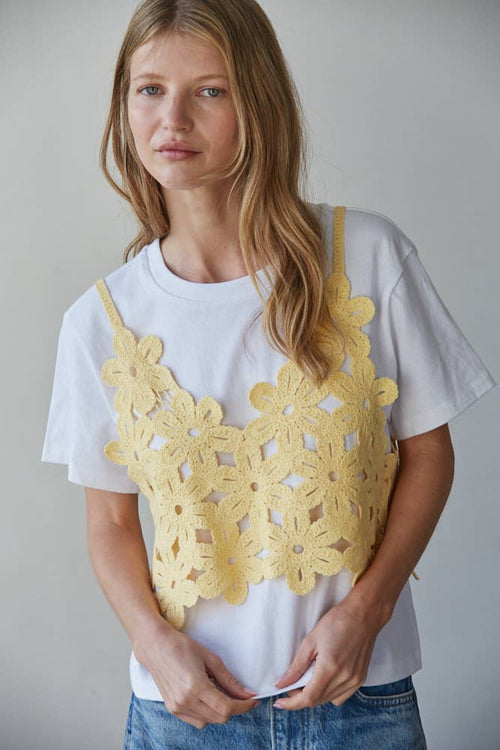 Woman wearing a white t-shirt with a yellow floral crochet top over it against a plain background