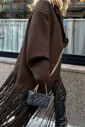 A person wearing an oversized dark brown coat with long fringe along the hem, carrying a small black quilted handbag with a gold chain strap. The photo is taken outdoors in front of a storefront window.
