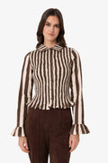 A woman modeling a brown and cream vertically striped long-sleeve blouse with a ruffled hem and cuffs, paired with dark brown pants, against a plain light gray background.