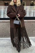 A woman wearing an oversized dark brown coat with a long fringe hem, styled with a black top, black fringe boots, and a black quilted handbag with a gold chain strap, standing on a city sidewalk.