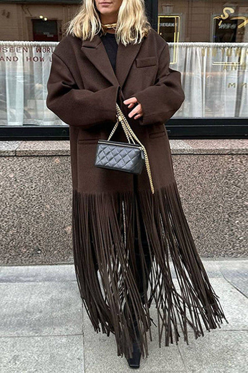 A woman wearing an oversized dark brown coat with a long fringe hem, styled with a black top, black fringe boots, and a black quilted handbag with a gold chain strap, standing on a city sidewalk.