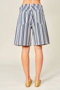 A person shown from the back wearing knee-length striped shorts with vertical navy, white, and beige stripes, paired with tan woven flat shoes against a plain light beige studio background.