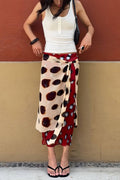 A woman wearing a beige wrap skirt with large black and dark red spots and a red side panel, paired with a white tank top and black flip-flops, standing against a two-toned wall.