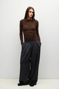 A woman modeling a brown long-sleeve ruched top and black wide-leg trousers against a plain light gray studio background.