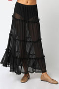 A black tiered sheer maxi skirt with ruffled trim, shown on a model wearing beige flat shoes against a light gray studio background.