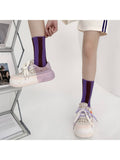 A person wearing purple and dark brown striped crew socks with chunky pastel sneakers, shown from the waist down in a bright studio setting.