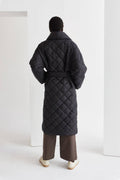 A model wearing a black quilted puffer wrap coat with a folded front collar and a self-belt with loops on the side. The coat has hidden pockets and is oversized for comfort.