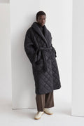 A model wearing a black quilted puffer wrap coat with a folded front collar and a self-belt with loops on the side. The coat has hidden pockets and is oversized for comfort.