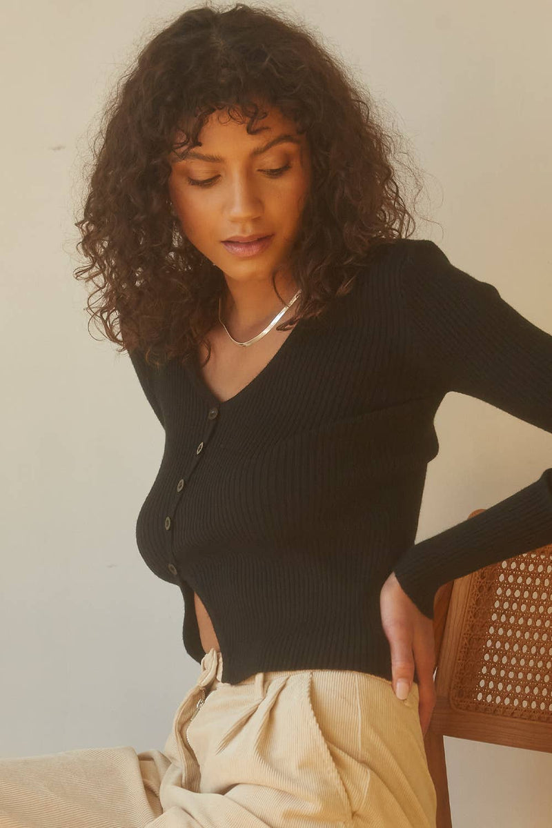 Woman wearing a black cardigan over a black top with beige pants, sitting on a wooden chair.