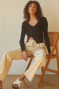 Woman sitting on a wooden chair wearing a black cardigan and beige pants.