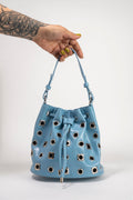 A light blue leather bucket bag with silver-tone metal studs and a drawstring closure, held by a tattooed hand with yellow nail polish against a plain white background.