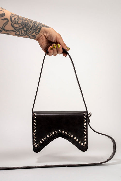 A hand with yellow nail polish and a tattooed arm holds a black leather shoulder bag with silver studs, against a plain light gray background.