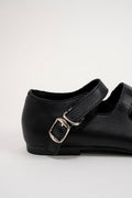 A close-up side view of a black leather Mary Jane shoe with a silver buckle, set against a plain light gray background.